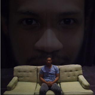 Main character Zeke, seated on a couch in front of a large projected image of his own face