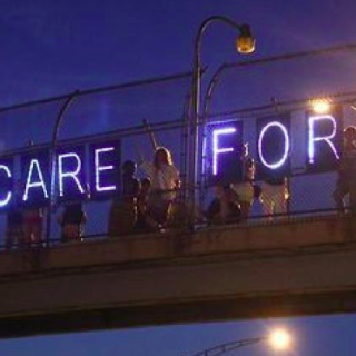 A row of people standing on a bridge at night, holding up illuminated letters that spell "HEALTH CARE FOR ALL"