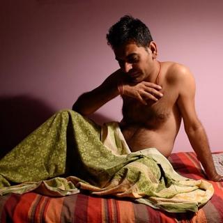 Photograph of a man with a bare chest reclining on a bed, draped in a green blanket.