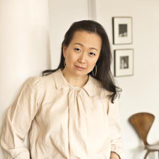 Photo of Min Jin Lee standing indoors, wearing hoop earrings, an off-white blouse and blue jeans