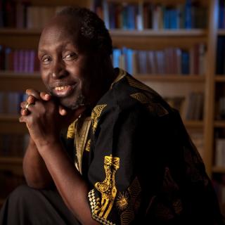Portrait of Ngũgĩ wa Thiong’o sitting in front of bookshelves with his hands clasped