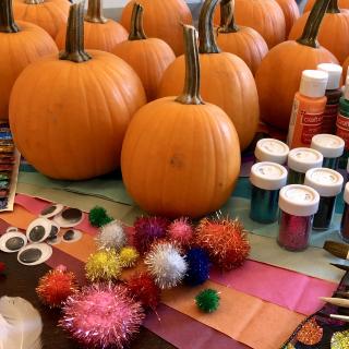 Pumpkins displayed with decorating supplies including paint, glitter, brushes, googly eyes, pom poms, tissue paper, sequins, feathers, and ribbon