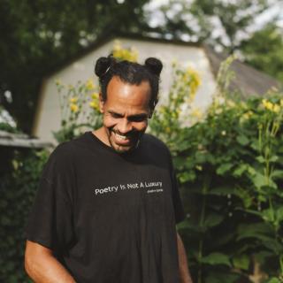 Ross Gay outside, surrounded by flowering plants and wearing a black shirt that says "'poetry is not a luxury' - Audre Lorde"