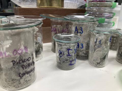Image shows 8 glass beakers of various sizes with writing on them and inside the beakers are grey colored sediment samples. In the background there are numerous petri dishes and a brown wooden box.