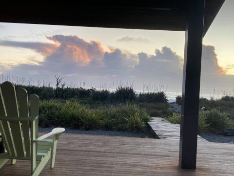 Image shows the a light pink cloud in the sky as the sun sets. The Pacific ocean is in the background and ahead of that are some plants. In the foreground there is a view of a porch and a light green lawn chair.