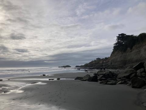 Image shows a beach that appears grey in the early morning light, a brown cliff face to the right, ocean to the left, and a light blue-grey sky with some clouds in the background 