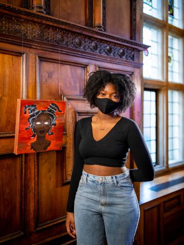 Ashanti stands next to her painting, Medusa. She wears a black long sleeve crop top and high rise blue jeans.