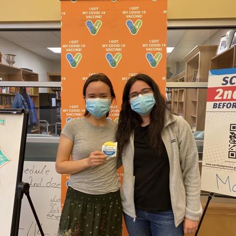 Posing with my friend and a sticker at the vaccine clinic. Masked and in front of an orange photo booth backdrop.