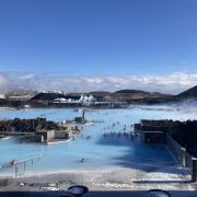 Image shows a bright blue pool with volcanic rocks around it and mountains in the background