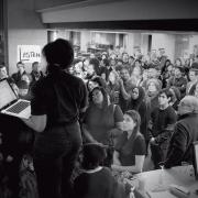 A black and white photo of a woman speaking to a large crowd inside a library