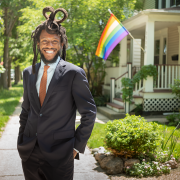 A Black man in a suit standing in a suburban neighborhood with a Pride flag in the background