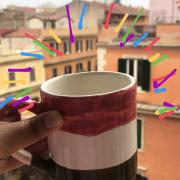 red, white, and green mug held against a view of warm-toned Italian buildings  